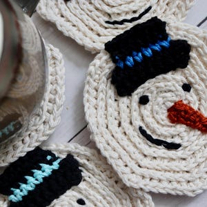 May include: Four crocheted snowman coasters in white with black eyes, black mouths, and black hats with blue trim. One coaster has a red nose.