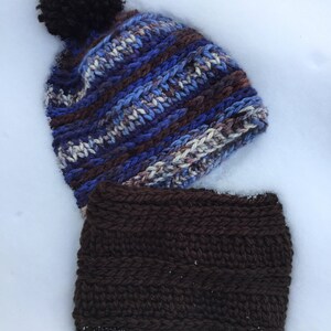 May include: A blue and brown striped knitted hat with a black pom pom and a matching brown knitted neck warmer. Both items are lying on a white snowy surface.