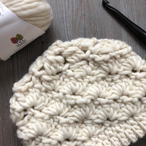 May include: A white crocheted hat made with chunky yarn. The ball of yarn and crochet hook are also visible in the image.