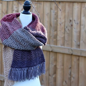 May include: A crocheted scarf with a chevron pattern in shades of brown, purple, and beige. The scarf has fringe on the ends.