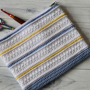 May include: A white crocheted zippered pouch with blue and yellow stripes. The pouch is lying on a white wooden surface.