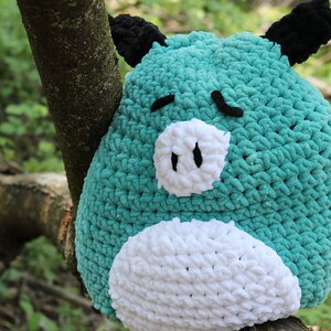 May include: A teal blue crocheted stuffed animal shaped like a pig with white crocheted details and black crocheted ears. The pig is sitting on a tree branch.