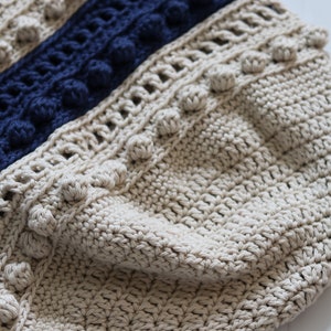 May include: Close-up of a beige crocheted bag with a blue and beige striped pattern. The bag has a textured surface and is decorated with small pom-poms.