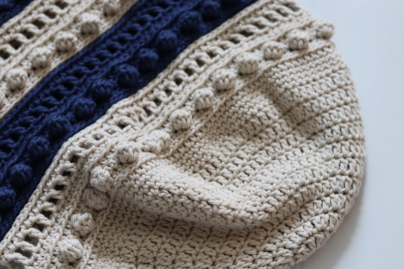 May include: Close-up of a beige crocheted bag with a blue and beige striped pattern. The bag has a textured surface and is decorated with small pom-poms.