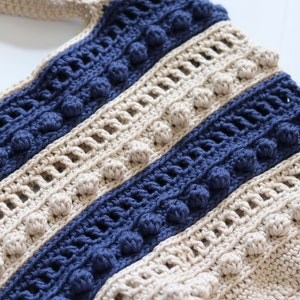 May include: A close-up of a crocheted bag with beige and blue stripes. The bag has a textured pattern and features small pom-poms along the edges of the stripes.