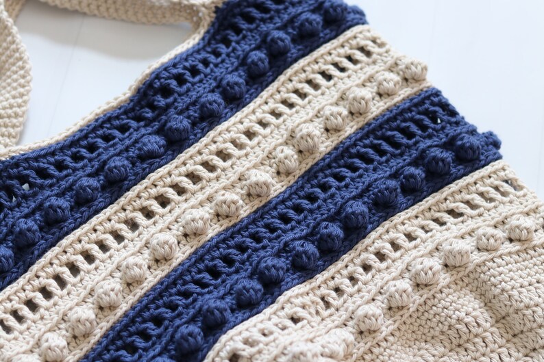 May include: A close-up of a crocheted bag with beige and blue stripes. The bag has a textured pattern and features small pom-poms along the edges of the stripes.
