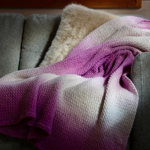 May include: A knitted throw blanket with a gradient color scheme of white, light pink, and deep purple. The blanket is draped over the arm of a gray sofa, with a fluffy beige pillow in the background. The blanket's texture is visible.