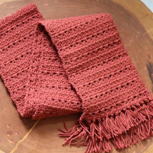 Peut inclure: Écharpe en crochet faite à la main, de couleur terre cuite, avec des franges. L'écharpe est faite d'un fil doux et présente un motif de point texturé avec de petits trous. Elle est présentée sur une surface en bois.