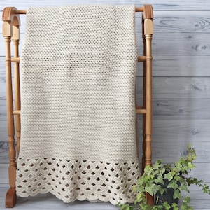 May include: A beige crocheted throw blanket with a delicate lace trim, displayed on a wooden stand against a white woodgrain background.