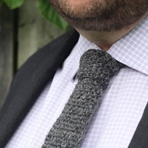 May include: A close-up of a man wearing a gray knitted tie with a white button-down shirt and a black blazer.