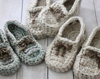 May include: Five pairs of crocheted slippers in beige and blue. The slippers have a loafer style with a brown crocheted bow detail.