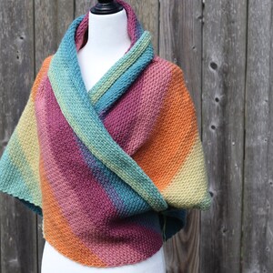 May include: A colorful crocheted shawl with a gradient of orange, pink, teal, and green. The shawl is draped over a white mannequin.