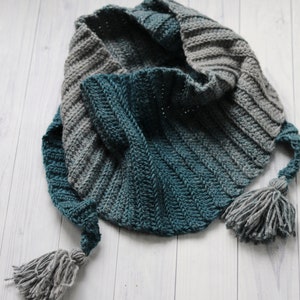 May include: A teal and gray crocheted scarf with tassels on each end. The scarf is folded in half and laid on a white wooden surface.