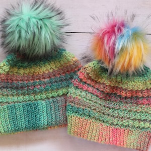 May include: Two colorful, handmade crochet beanies with faux fur pom-poms. One beanie has a mint green pom-pom, the other a rainbow pom-pom. The hats feature a mix of green, yellow, orange, and pink yarn, perfect for cold weather.