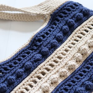 May include: A crocheted tote bag with a striped pattern in beige and navy blue. The bag has a wide, flat handle and features a textured stitch with small, raised bobbles.