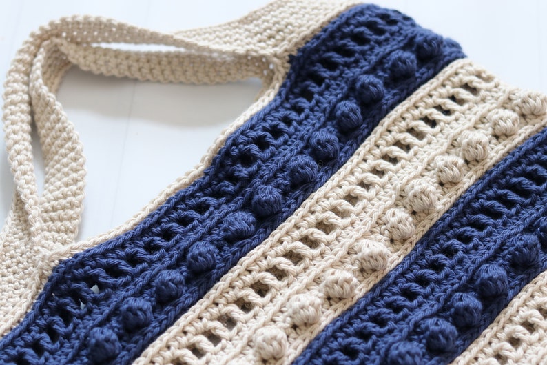 May include: A crocheted tote bag with a striped pattern in beige and navy blue. The bag has a wide, flat handle and features a textured stitch with small, raised bobbles.