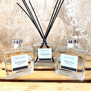 May include: Three clear glass bottles with black labels and black and white striped designs. The bottles are filled with a clear liquid and have black reeds sticking out of the top. The labels read "Saltgrass Candles", "Lush Linen", "White Birch", "Black Sea", and "Reed Diffuser".