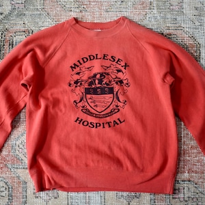 May include: A coral-coloured sweatshirt with long sleeves. The front displays a black crest with the words "MIDDLESEX HOSPITAL" in a serif font. The sweatshirt is made of a soft material and has a crew neck.