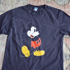 May include: A dark charcoal gray short-sleeve t-shirt featuring a graphic print of Mickey Mouse on the front. The character is depicted in a classic pose with a white face, black ears, red shorts, and large yellow shoes. A small tag is at the neck.