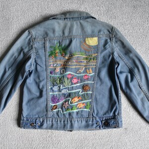 May include: A blue denim jacket with an embroidered scene of a beach with palm trees, waves, and fish. The embroidery is colorful and detailed.