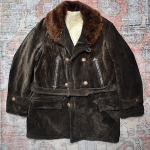 Puede incluir: Un abrigo vintage de pana marrón con cuello de piel sintética y cinturón en la cintura. El abrigo cruzado presenta botones grandes, bolsillos y detalles de cuero. Una prenda de abrigo clásica.