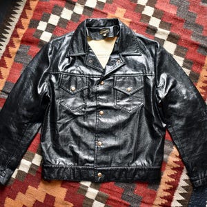 May include: A black jacket with a glossy finish, featuring a pointed collar, long sleeves, and a button-front closure. The garment includes two chest pockets with buttoned flaps and a tan interior lining visible at the neckline and collar area.