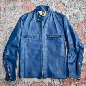 Puede incluir: Chaqueta de cuero azul con cuello alzado, dos bolsillos en el pecho con cremallera y puños con cremallera. La chaqueta tiene cierre de cremallera completa y forro amarillo visible en el cuello y los puños.