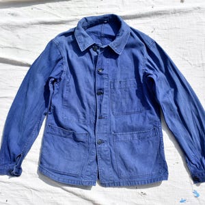 May include: A blue work jacket featuring a standard collar, long sleeves with buttoned cuffs, and a front button closure. The garment includes one chest pocket on the left side and two larger patch pockets positioned at the lower front section.
