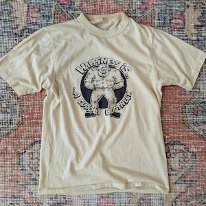 May include: A beige t-shirt with a graphic of a strong figure inside a circle. The text reads "Happiness is: A Cuddly Bootneck". The shirt has short sleeves and a round neck. The t-shirt is made of a soft material.