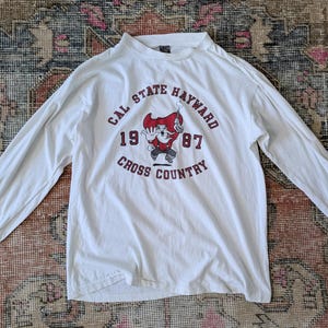 Könnte beinhalten: Weißes Langarmshirt mit Rundhalsausschnitt. Das Shirt hat einen Aufdruck mit dem Text "CAL STATE HAYWARD 1987 CROSS COUNTRY" und einem Cartoon-Maskottchen in Rot, Weiß und Schwarz. Das Shirt liegt flach auf einem gemusterten Teppich.