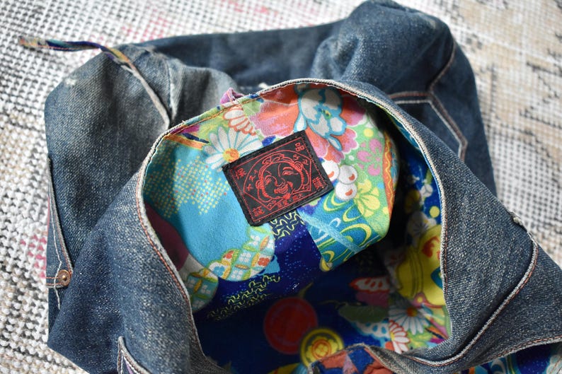 Puede incluir: Un bolso tote de mezclilla con un interior estampado floral colorido. El interior del bolso presenta una vibrante mezcla de dise&ntilde;os florales azules, verdes y amarillos. Una etiqueta negra con texto rojo es visible dentro del bolso.
