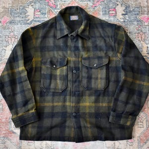 May include: A green and black checked shirt with two chest pockets and a button-down front. The shirt has a collared neckline and long sleeves. The fabric appears to be a thick wool or wool blend.