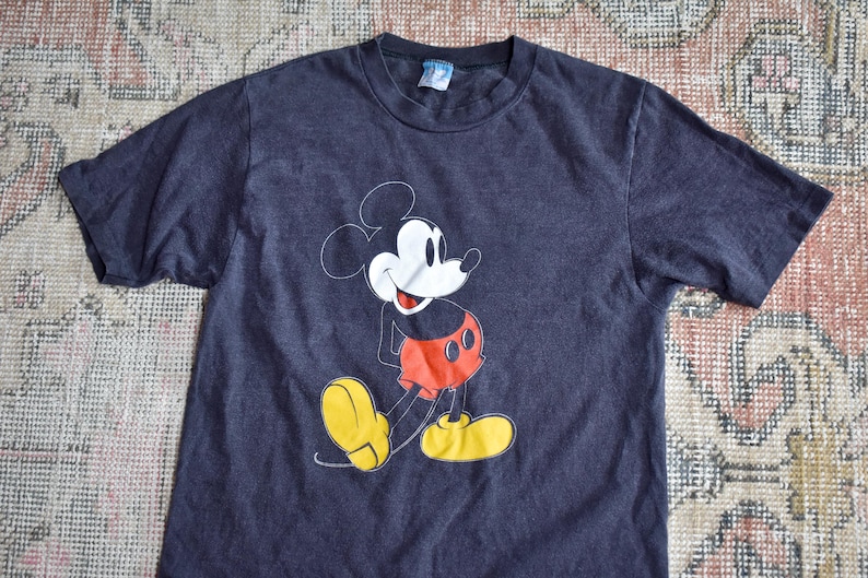 May include: A dark charcoal gray short-sleeve t-shirt featuring a graphic print of Mickey Mouse on the front. The character is depicted in a classic pose with a white face, black ears, red shorts, and large yellow shoes. A small tag is at the neck.