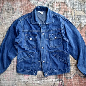 May include: A vintage denim jacket in a medium blue wash. The jacket has a collared neckline, button-down closure, and multiple pockets. The long sleeves are cuffed at the wrists. The jacket is laid flat on a patterned rug.