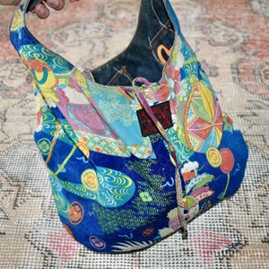 Puede incluir: Bolso de hombro azul estampado con interior de mezclilla. El bolso presenta un dise&ntilde;o colorido con motivos de inspiraci&oacute;n japonesa en tonos azules, amarillos, rosas y verdes. Tiene cierre de cord&oacute;n y un asa.