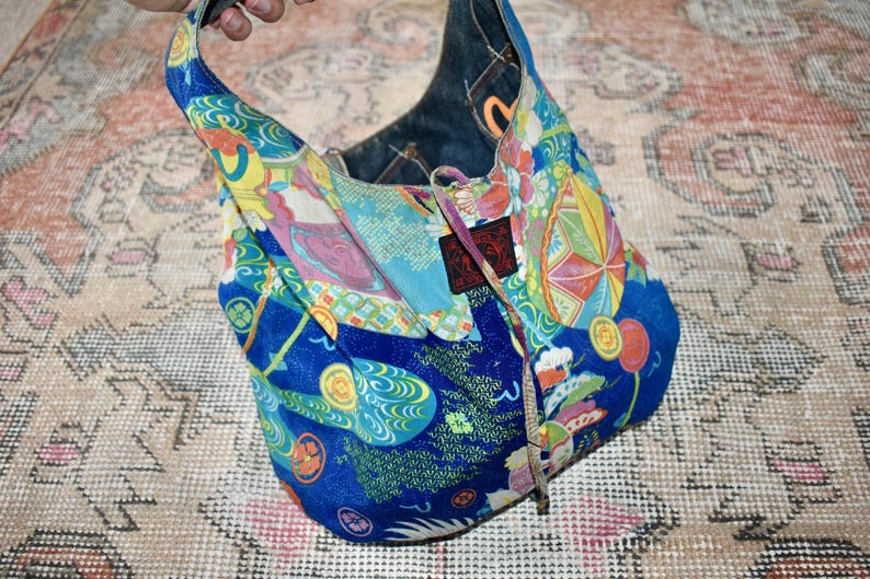 Puede incluir: Bolso de hombro azul estampado con interior de mezclilla. El bolso presenta un dise&ntilde;o colorido con motivos de inspiraci&oacute;n japonesa en tonos azules, amarillos, rosas y verdes. Tiene cierre de cord&oacute;n y un asa.