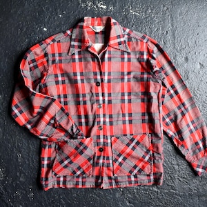 May include: A long-sleeve button-down shirt featuring a red, black, and white plaid pattern. The shirt includes a pointed collar, a front button closure, and two square patch pockets positioned at the lower front hem with a diagonal plaid design.