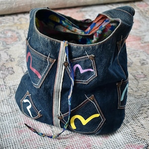 Puede incluir: Un bolso tote de mezclilla azul oscuro con múltiples bolsillos, cada uno adornado con diseños de ondas estilizadas y coloridas. El bolso presenta un cierre de cremallera y un interior estampado. Una cuerda decorativa cuelga del bolso.