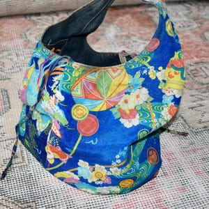 Puede incluir: Un bolso de mano de tela azul con un colorido estampado floral y geom&eacute;trico. El bolso tiene un asa de mezclilla y un cierre de cord&oacute;n. El dise&ntilde;o incluye flores, abanicos y motivos de inspiraci&oacute;n japonesa.