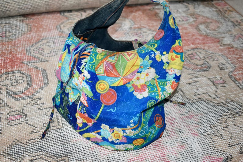 Puede incluir: Un bolso de mano de tela azul con un colorido estampado floral y geom&eacute;trico. El bolso tiene un asa de mezclilla y un cierre de cord&oacute;n. El dise&ntilde;o incluye flores, abanicos y motivos de inspiraci&oacute;n japonesa.