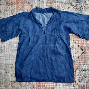 Puede incluir: Una blusa de mezclilla azul con mangas cortas y cuello en V. La blusa tiene un corte holgado y pliegues en la cintura. La tela parece ser una mezclilla de peso medio. La blusa está tendida sobre una alfombra estampada.