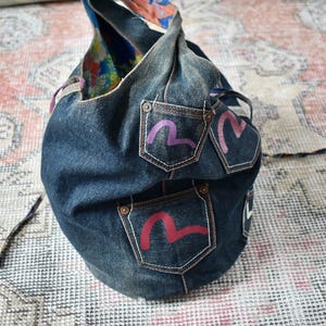 Puede incluir: Un bolso de mano de mezclilla azul oscuro con tres bolsillos, cada uno con un dise&ntilde;o estilizado y colorido. El bolso tiene forma redondeada y un asa. El forro interior es de tela estampada colorida.