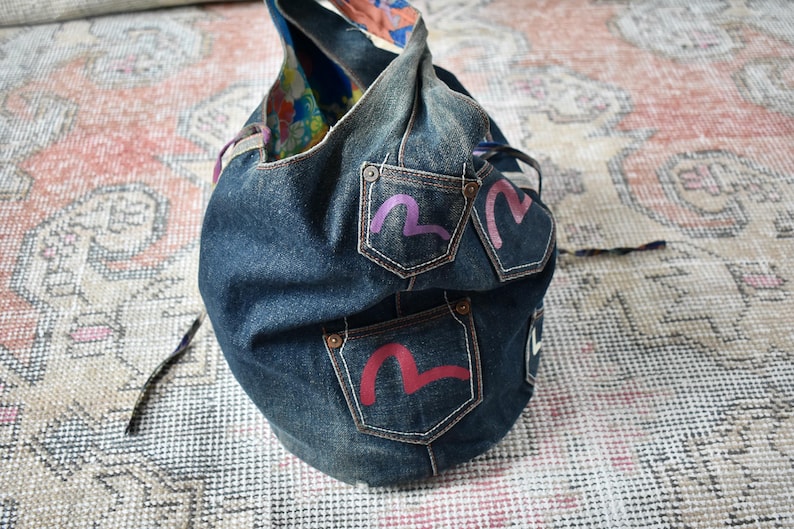 Puede incluir: Un bolso de mano de mezclilla azul oscuro con tres bolsillos, cada uno con un dise&ntilde;o estilizado y colorido. El bolso tiene forma redondeada y un asa. El forro interior es de tela estampada colorida.
