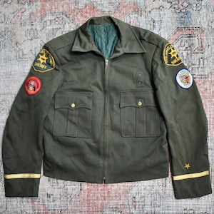 以下が含まれることがあります： オリーブグリーンのジャケット。ジッパー開閉、胸ポケット2つ、下ポケット2つ。袖口にゴールドのアクセント、袖に保安官の星などのワッペン付き。ヴィンテージスタイルのジャケット。