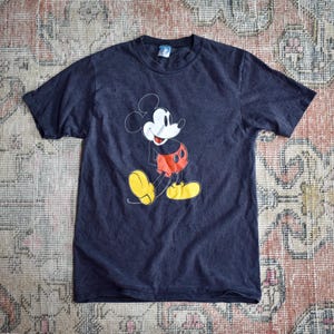 May include: A dark charcoal gray short-sleeve t-shirt featuring a graphic print of Mickey Mouse on the front. The character is depicted with a white face, red shorts, and yellow shoes, rendered in a classic line-art style on the chest area.