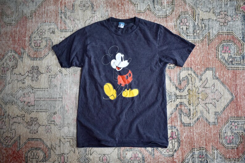 May include: A dark charcoal gray short-sleeve t-shirt featuring a graphic print of Mickey Mouse on the front. The character is depicted with a white face, red shorts, and yellow shoes, rendered in a classic line-art style on the chest area.