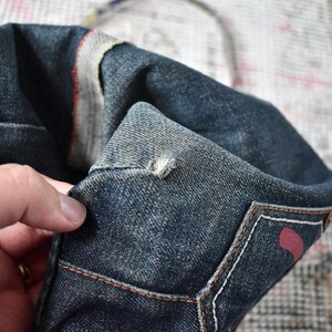 Puede incluir: Primer plano de un bolso tote de mezclilla azul oscuro. El bolso tiene un aspecto desgastado con costuras visibles y un acento rojo. El bolso est&aacute; sostenido por una mano, mostrando la textura y los detalles del material de mezclilla.