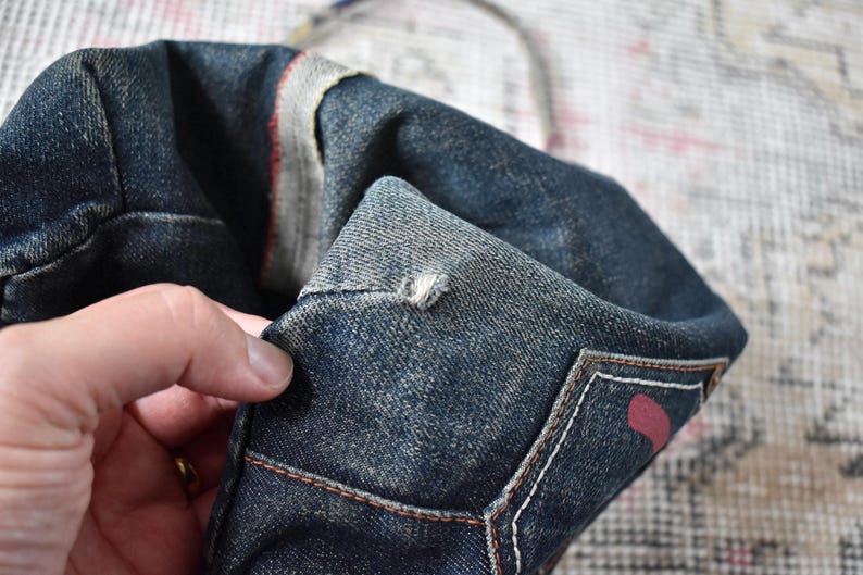 Puede incluir: Primer plano de un bolso tote de mezclilla azul oscuro. El bolso tiene un aspecto desgastado con costuras visibles y un acento rojo. El bolso est&aacute; sostenido por una mano, mostrando la textura y los detalles del material de mezclilla.