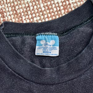 May include: A black crew neck t-shirt featuring a blue and white rectangular tag at the inner collar. The tag displays the Disney logo, the text Character Fashions, Walt Disney Productions, Made in USA, and the size Medium with care instructions.