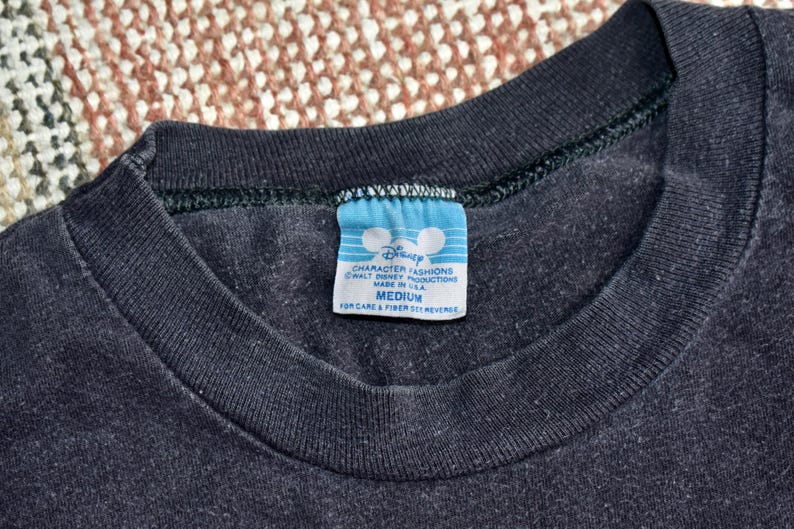 May include: A black crew neck t-shirt featuring a blue and white rectangular tag at the inner collar. The tag displays the Disney logo, the text Character Fashions, Walt Disney Productions, Made in USA, and the size Medium with care instructions.